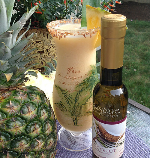 A gourmet piña colada cocktail made with Gustare Coconut Balsamic Vinegar, served in a frosted glass with a toasted coconut rim and fresh pineapple garnish