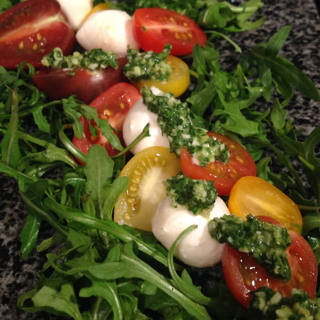 Gourmet caprese salad with colorful heirloom tomatoes, fresh mozzarella, and homemade arugula basil pesto served on peppery greens, showcasing our premium olive oils and Italian culinary ingredients.